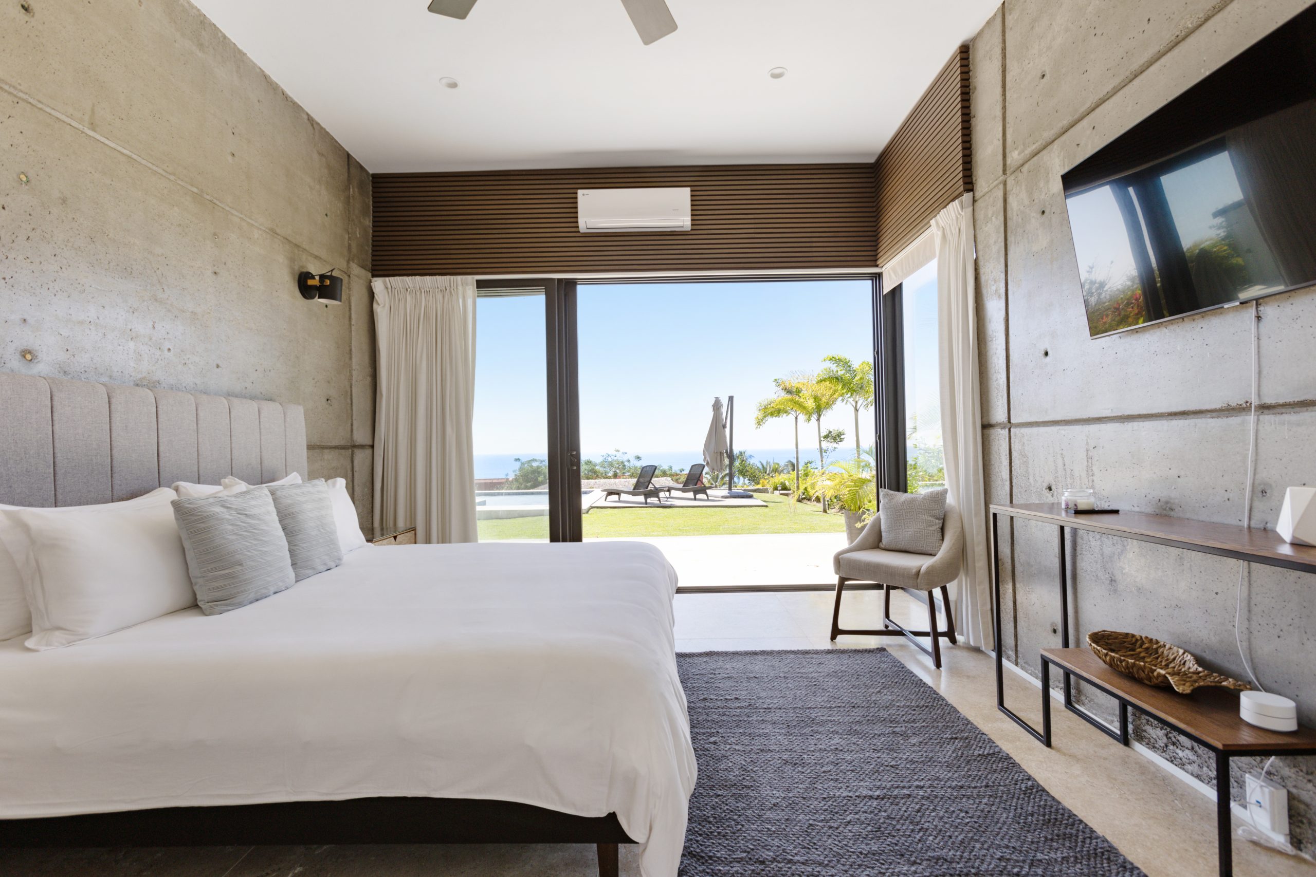 edroom with panoramic ocean views through large corner windows, light-colored curtains, and a cozy chair with a blanket.