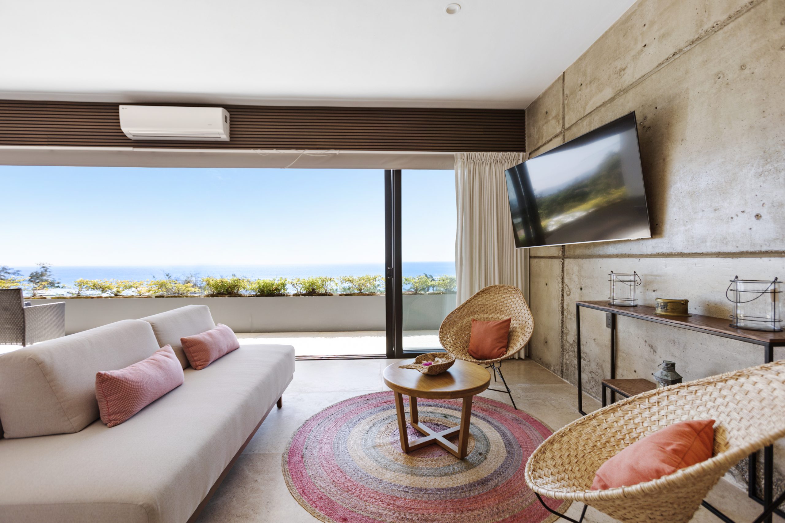 Modern living room with concrete accent wall, large TV, yellow sofa, gray armchairs, and glass doors opening to a pool view.
