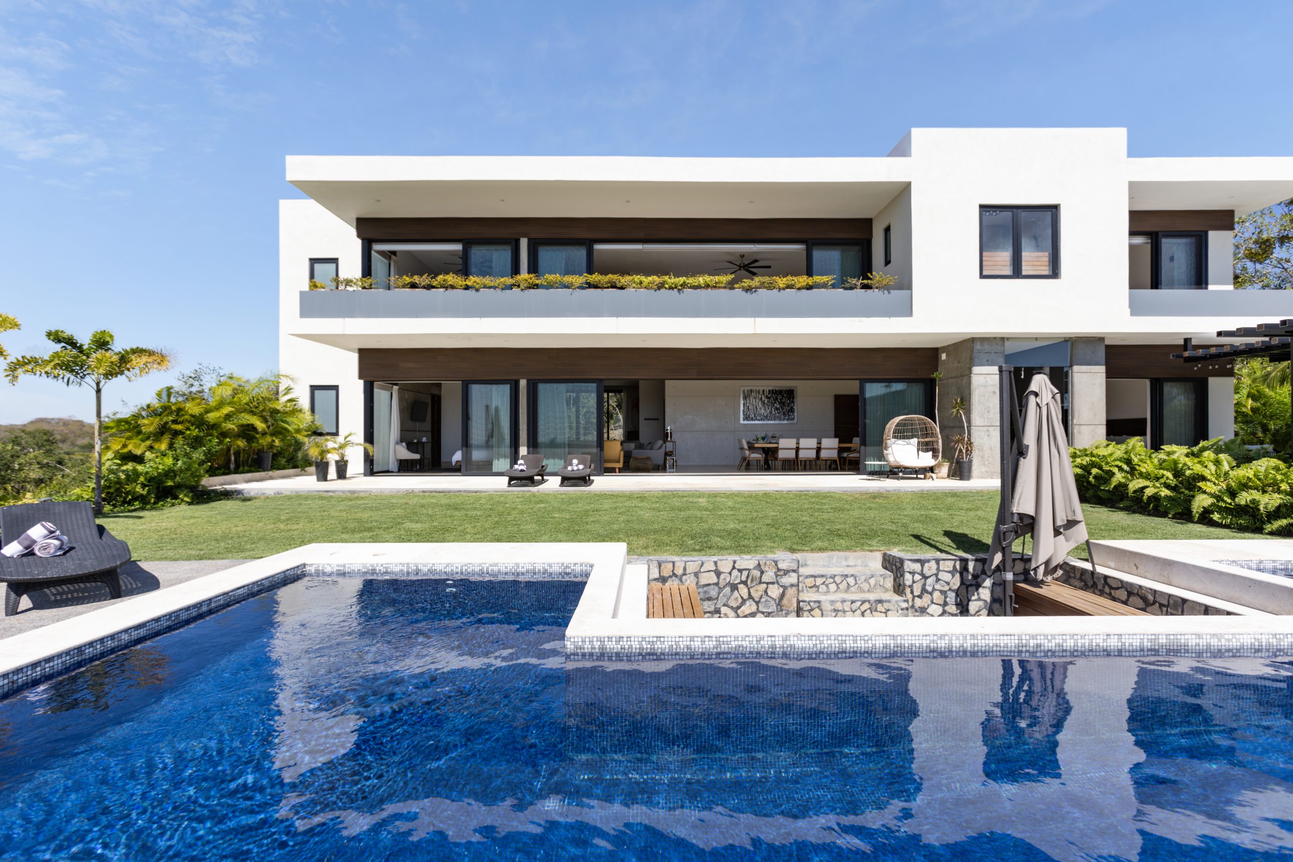 Modern two-story coastal home with white walls, large glass windows, and an infinity pool surrounded by lush greenery under a clear blue sky