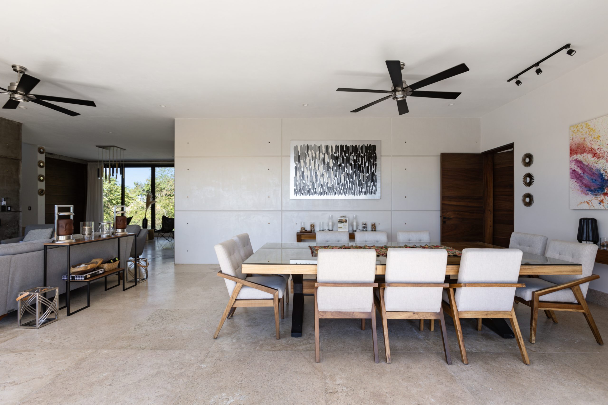 Spacious dining area in a contemporary home with a wooden table, upholstered chairs, ceiling fans, and minimalist artwork on the wall.