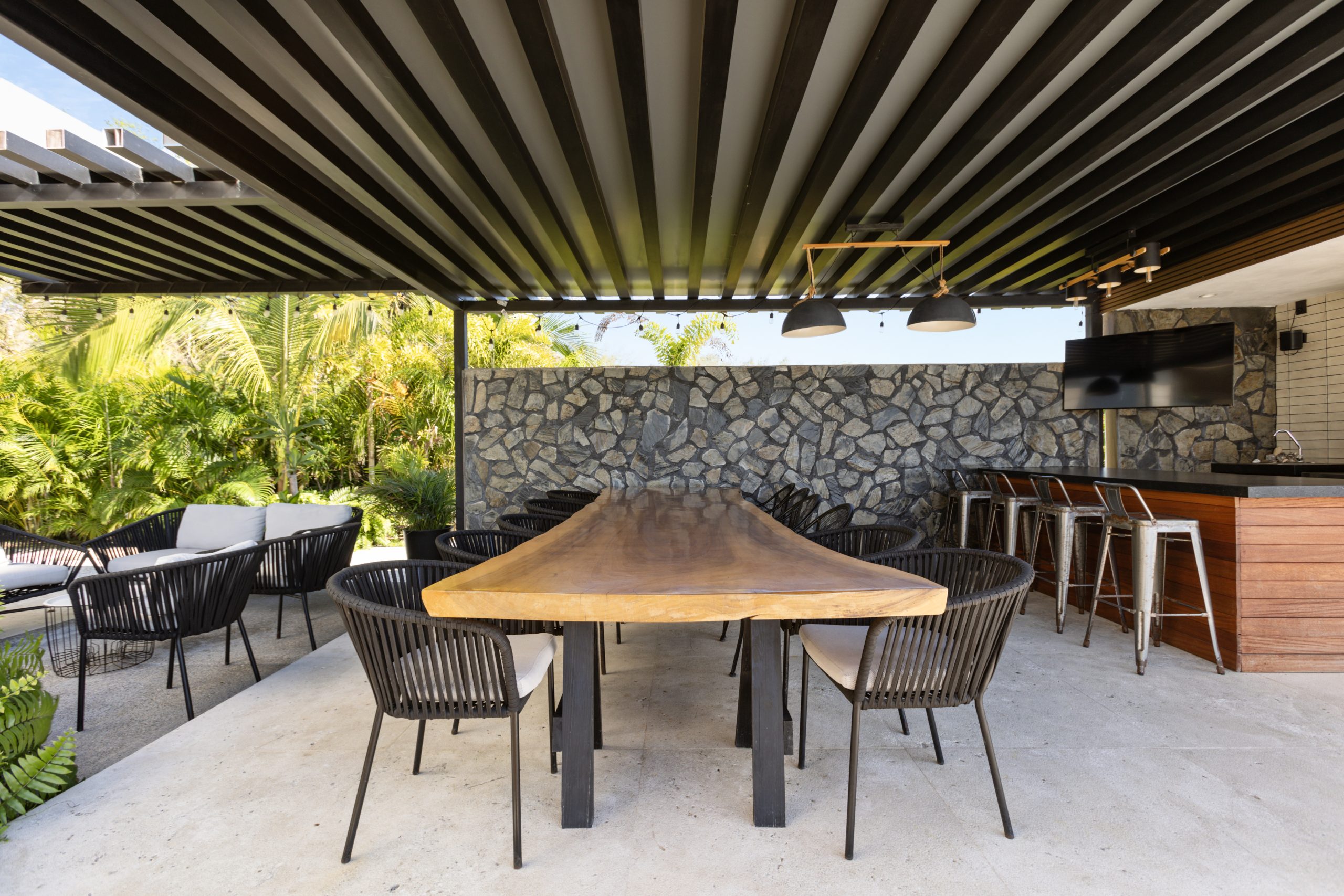 Outdoor covered dining area with a long wooden table, black chairs, pendant lights, and a stone wall near a tropical garden.