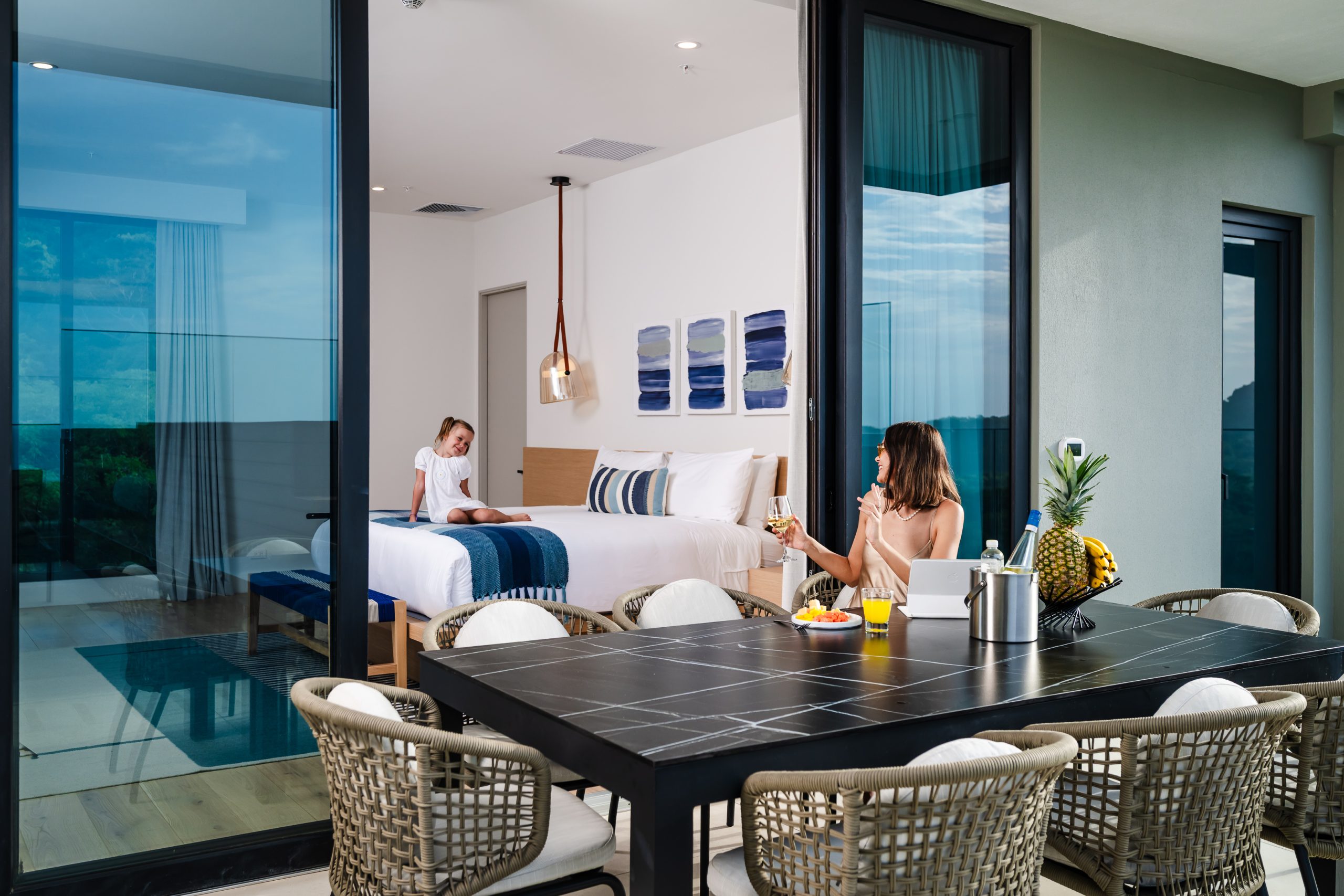Child playfully jumping on the bed while the mother relaxes on the outdoor terrace of a luxury Reserva Conchal penthouse, overlooking the lush Costa Rican landscape.